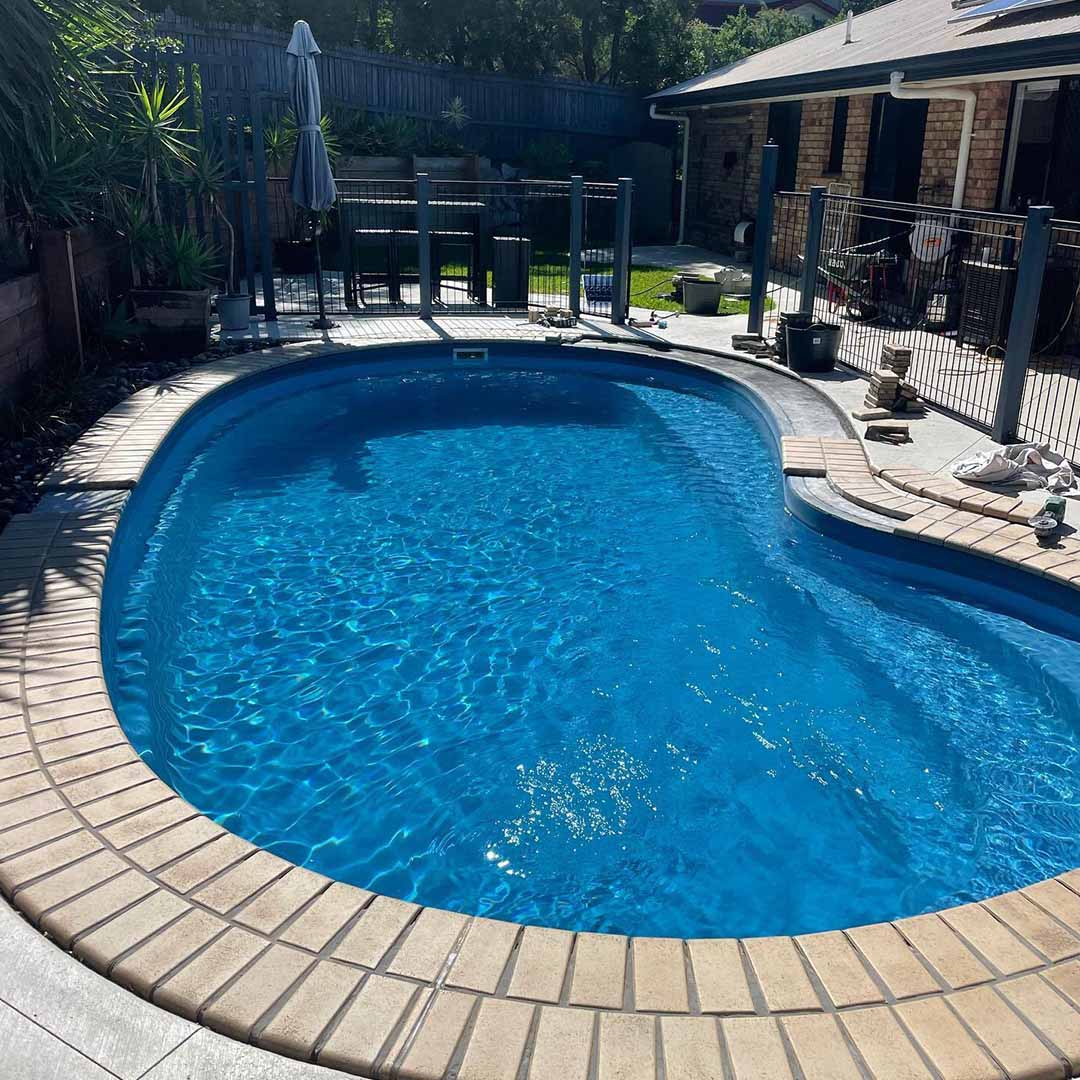 Kidney-shaped pool with brick-style coping and pale paving in a fenced yard