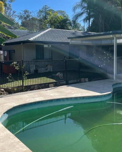 Pale stone surround around a pool with green water in a fenced backyard