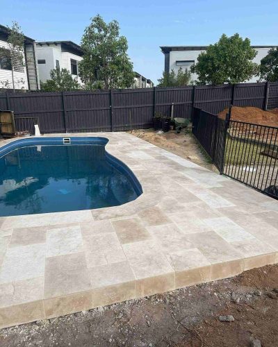 Pale stone pavers surrounding a kidney-shaped pool in a fenced backyard